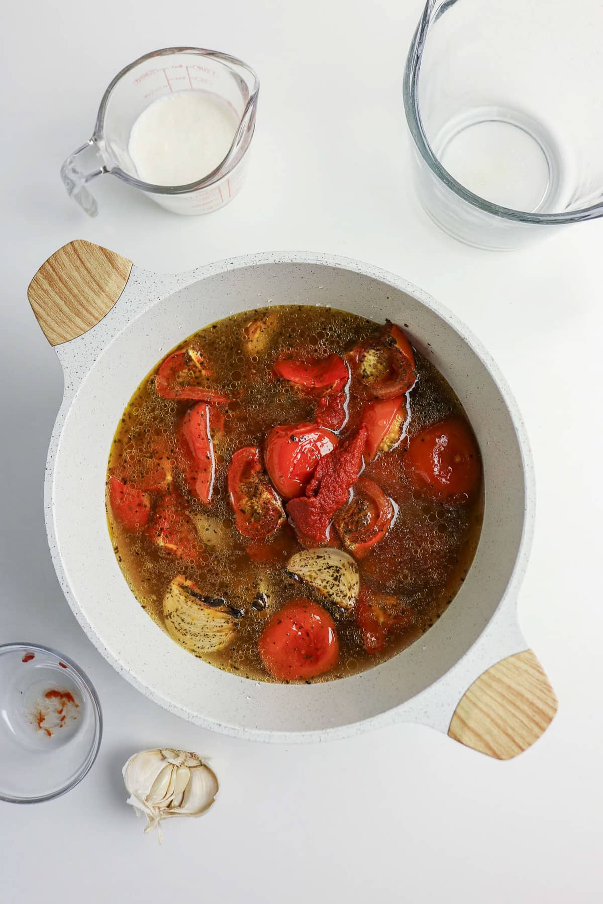 White pot with tomatoes, onion, and broth. Nearby are empty glass bowls, a garlic bulb, and a measuring cup of milk.
