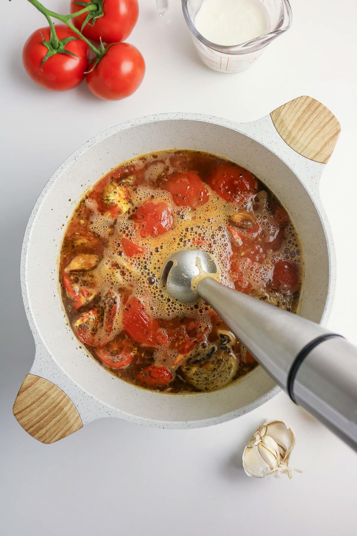 An immersion blender blends tomato soup in a pot, with fresh tomatoes and garlic nearby on the counter.