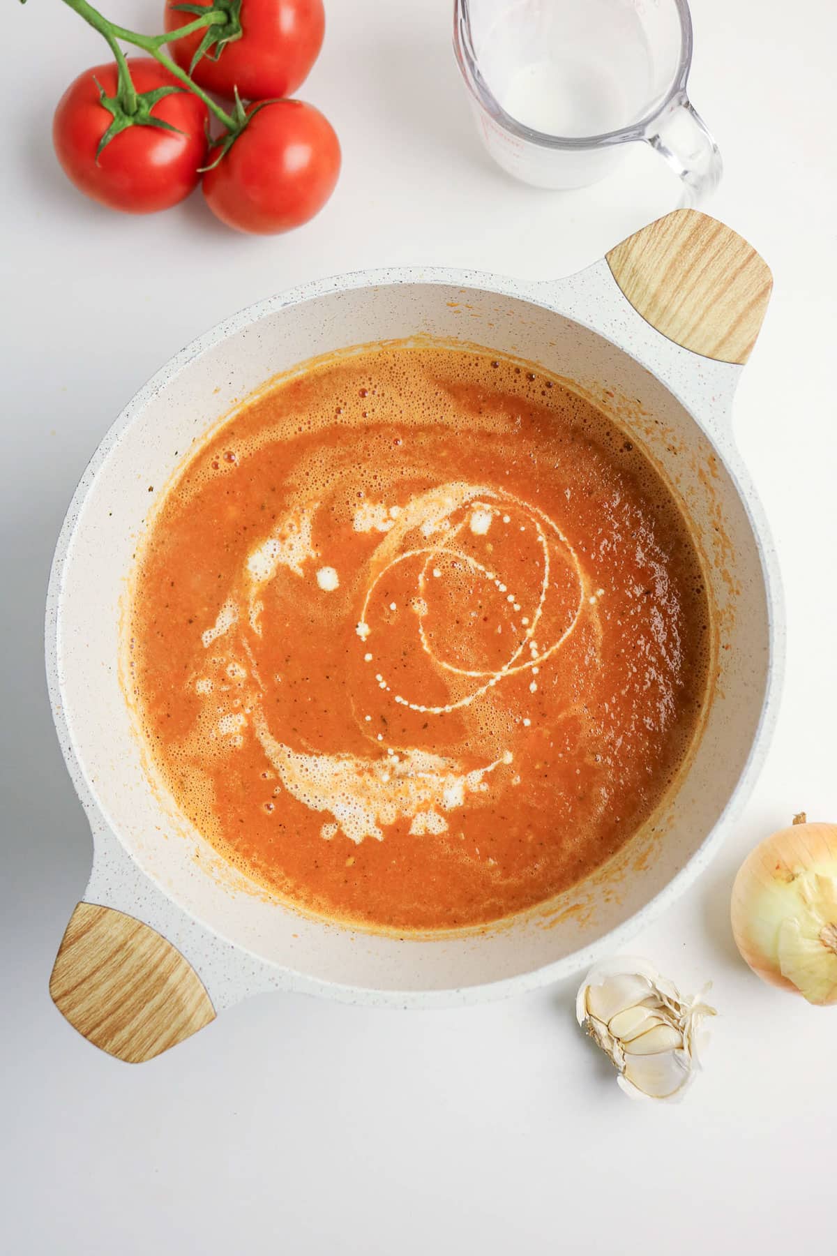 A pot of creamy tomato soup, with a swirl of cream, next to tomatoes, garlic, onion, and a measuring cup.