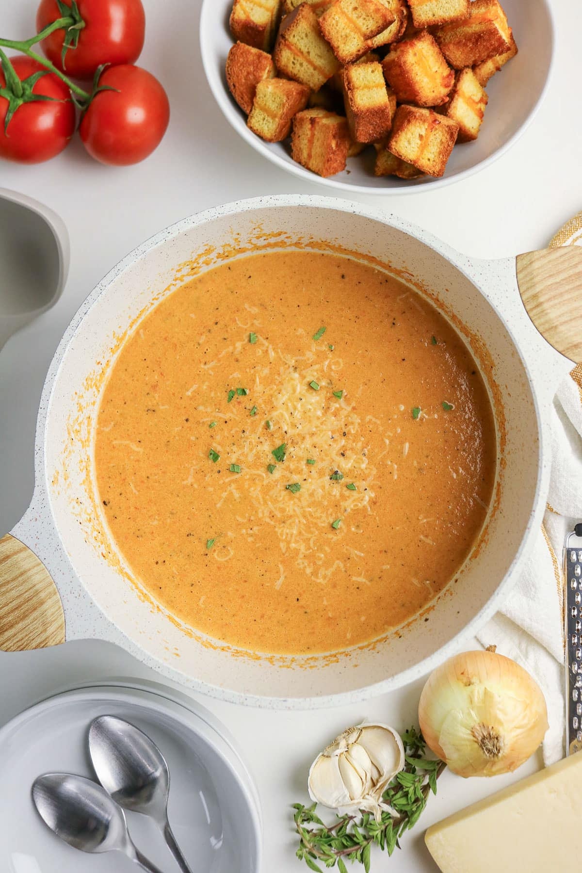 Creamy tomato soup in a pot, topped with herbs, surrounded by croutons, tomatoes, onion, garlic, and spoons.