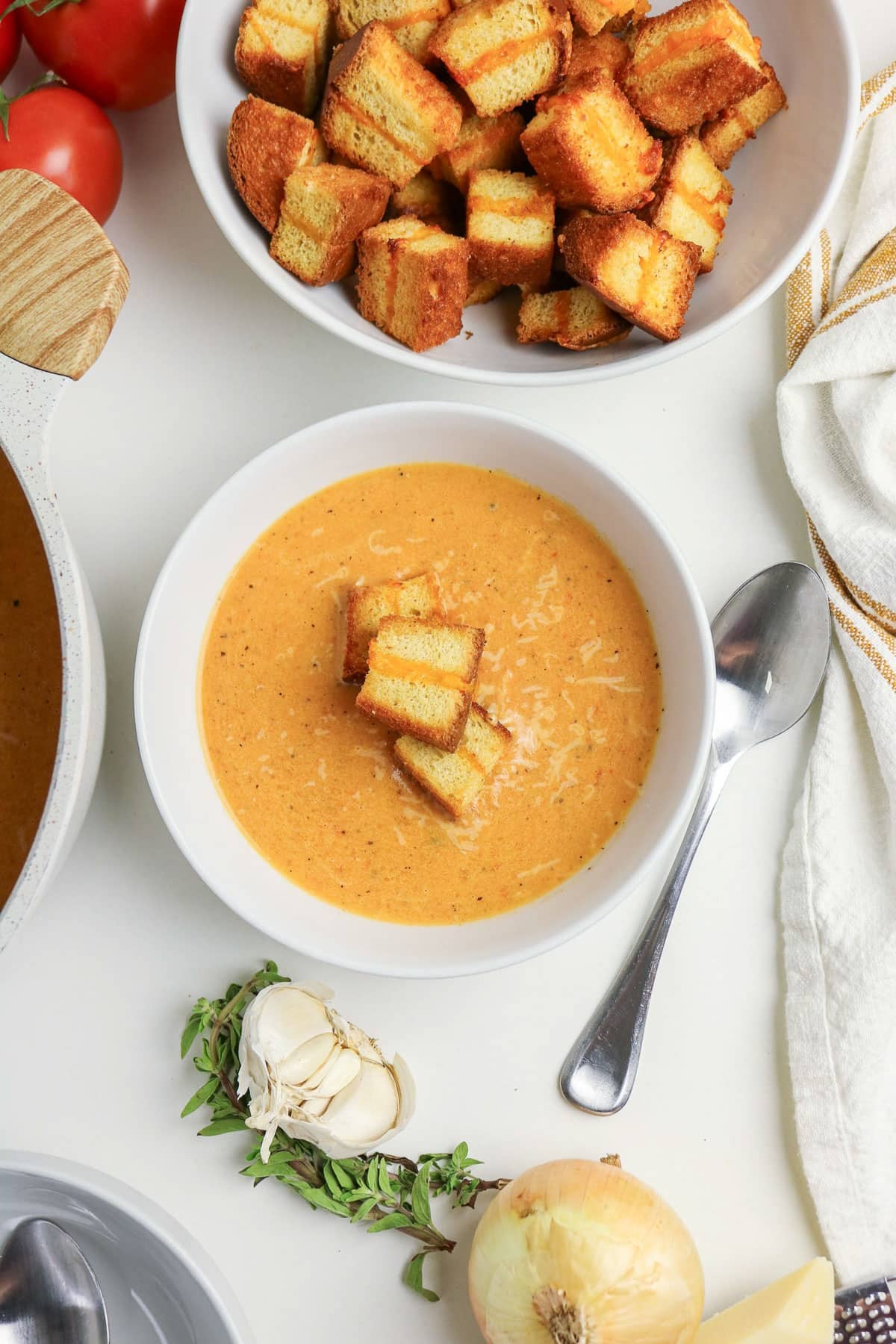 Bowl of creamy tomato soup topped with croutons, next to a bowl of croutons, herbs, garlic, and an onion.