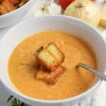 Creamy tomato soup in a white bowl topped with croutons, with a spoon and vegetables in the background.