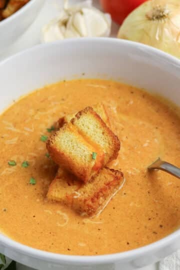 Creamy tomato soup in a white bowl topped with croutons, with a spoon and vegetables in the background.