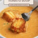A bowl of roasted tomato soup topped with savory grilled cheese croutons, served with a spoon on the side.