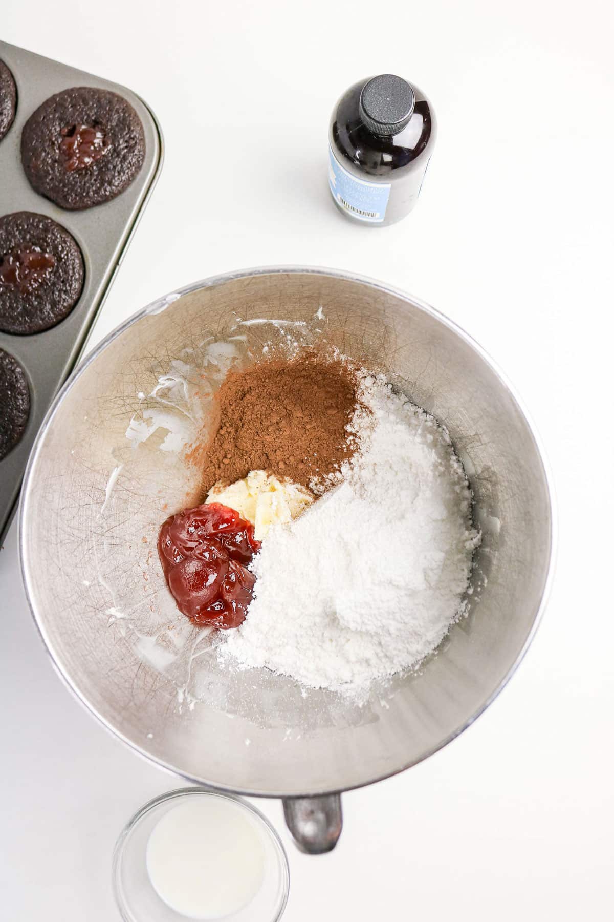 A mixing bowl with cocoa, sugar, butter, and jam, with milk, vanilla, and chocolate cupcakes nearby.