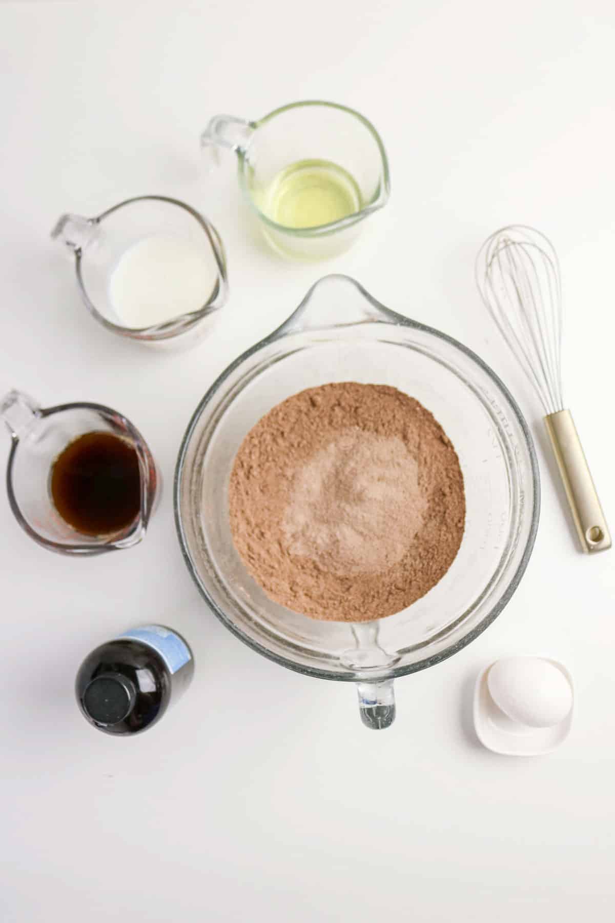 A bowl of cocoa powder mix, with milk, oil, vanilla, an egg, a whisk, and vanilla extract on a white table.