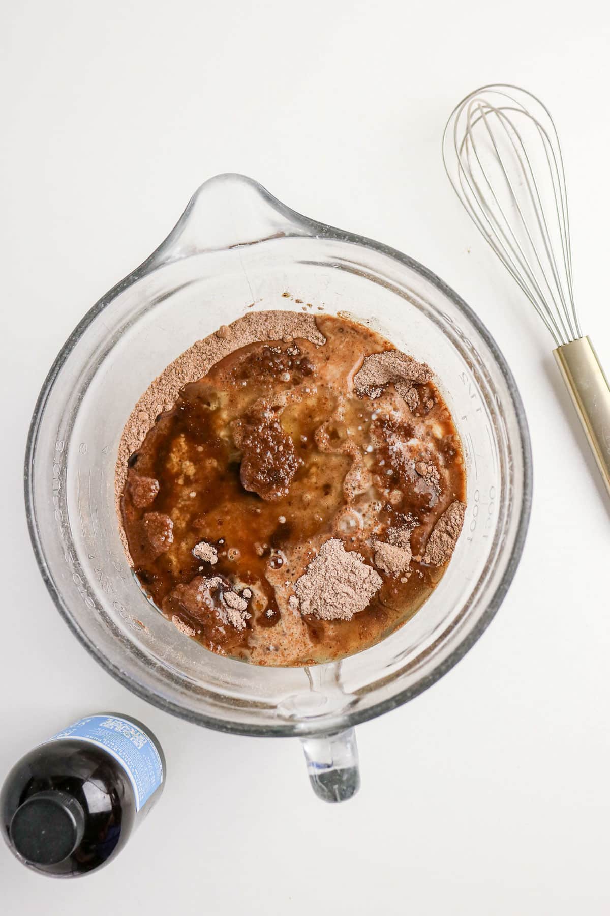 A glass bowl with cocoa powder and liquid ingredients, a whisk, and a bottle of vanilla extract nearby.
