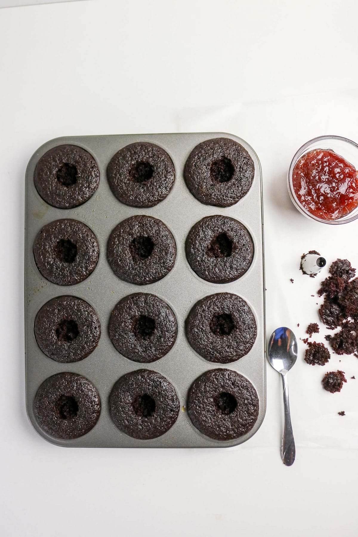 A muffin tin with 12 chocolate cupcakes, some with centers removed, next to jam, crumbs, and a spoon.