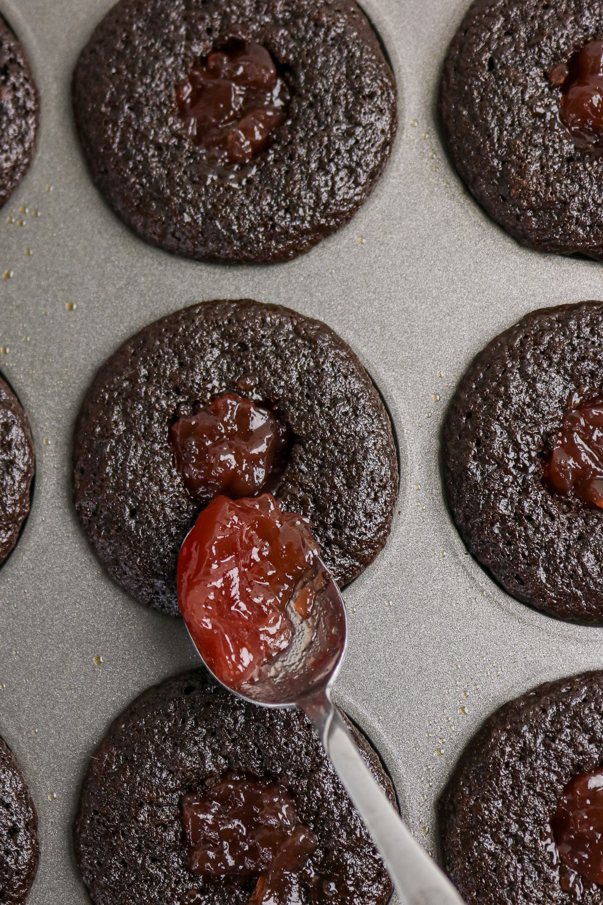 A spoon adds red jam to the center of baked chocolate cookies in a muffin tin.