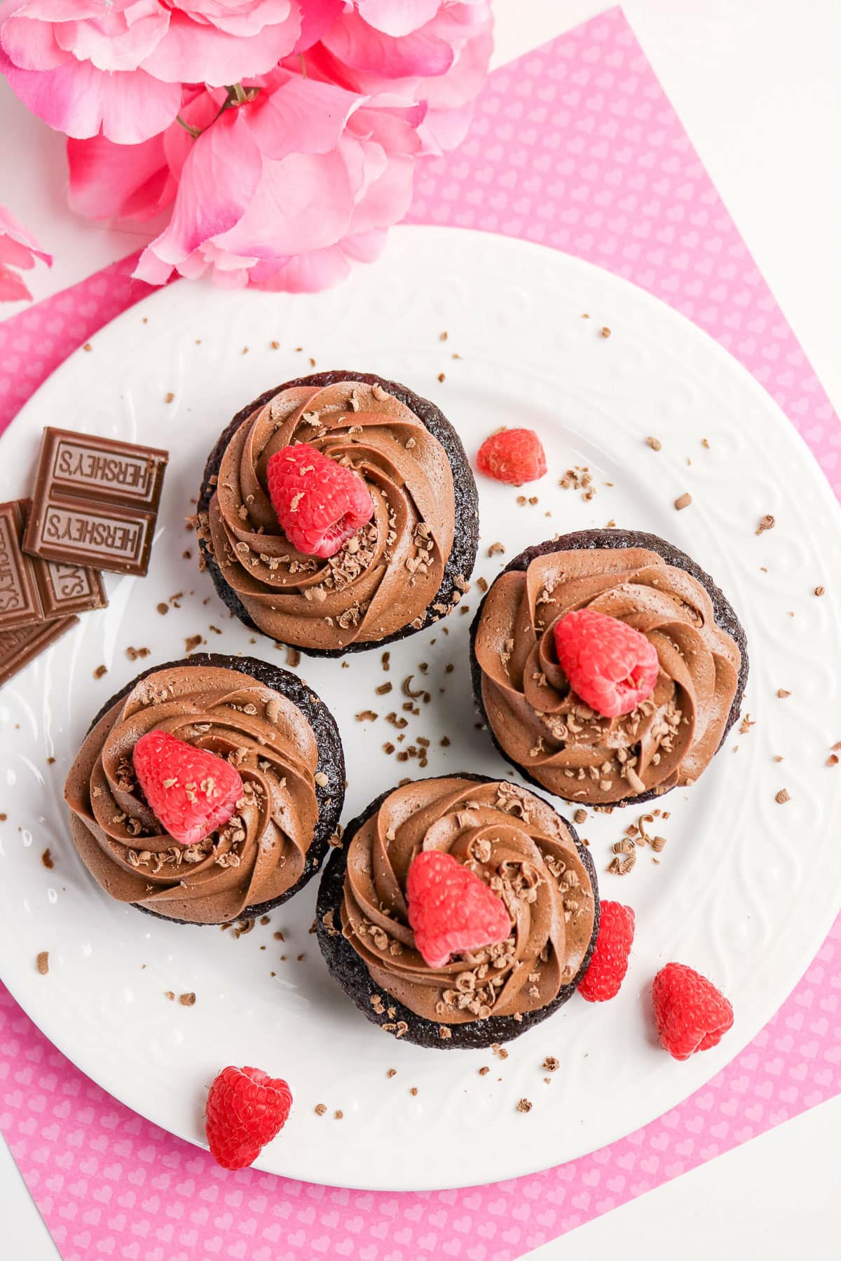 Four chocolate cupcakes with chocolate frosting and raspberries on top, on a white plate with chocolate pieces.