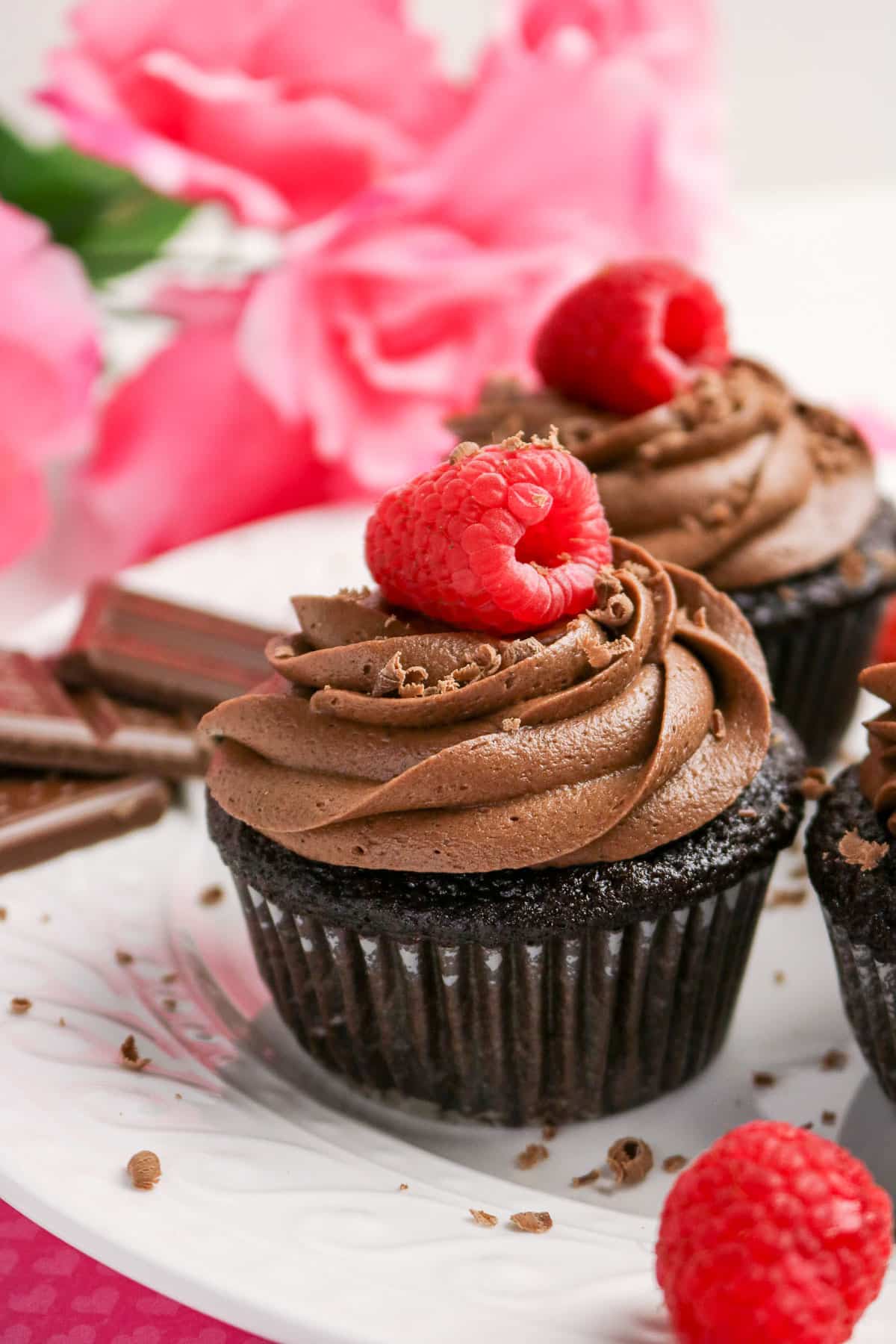 Chocolate cupcake with chocolate frosting, topped with a fresh raspberry, on a white plate with pink flowers.