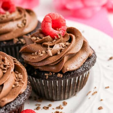 Chocolate cupcake with swirled frosting, topped with a raspberry and chocolate shavings on a white plate.