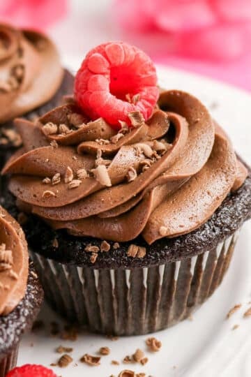 Chocolate cupcake with swirled frosting, topped with a raspberry and chocolate shavings on a white plate.