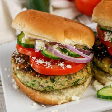 Turkey burger with tomato, cucumber, red onion, and crumbled cheese on a bun, served on a white plate.