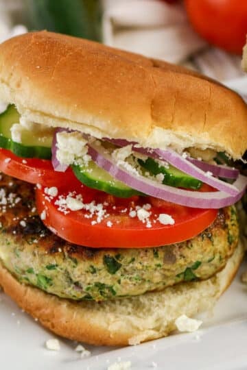 Turkey burger with tomato, cucumber, red onion, and crumbled cheese on a bun, served on a white plate.