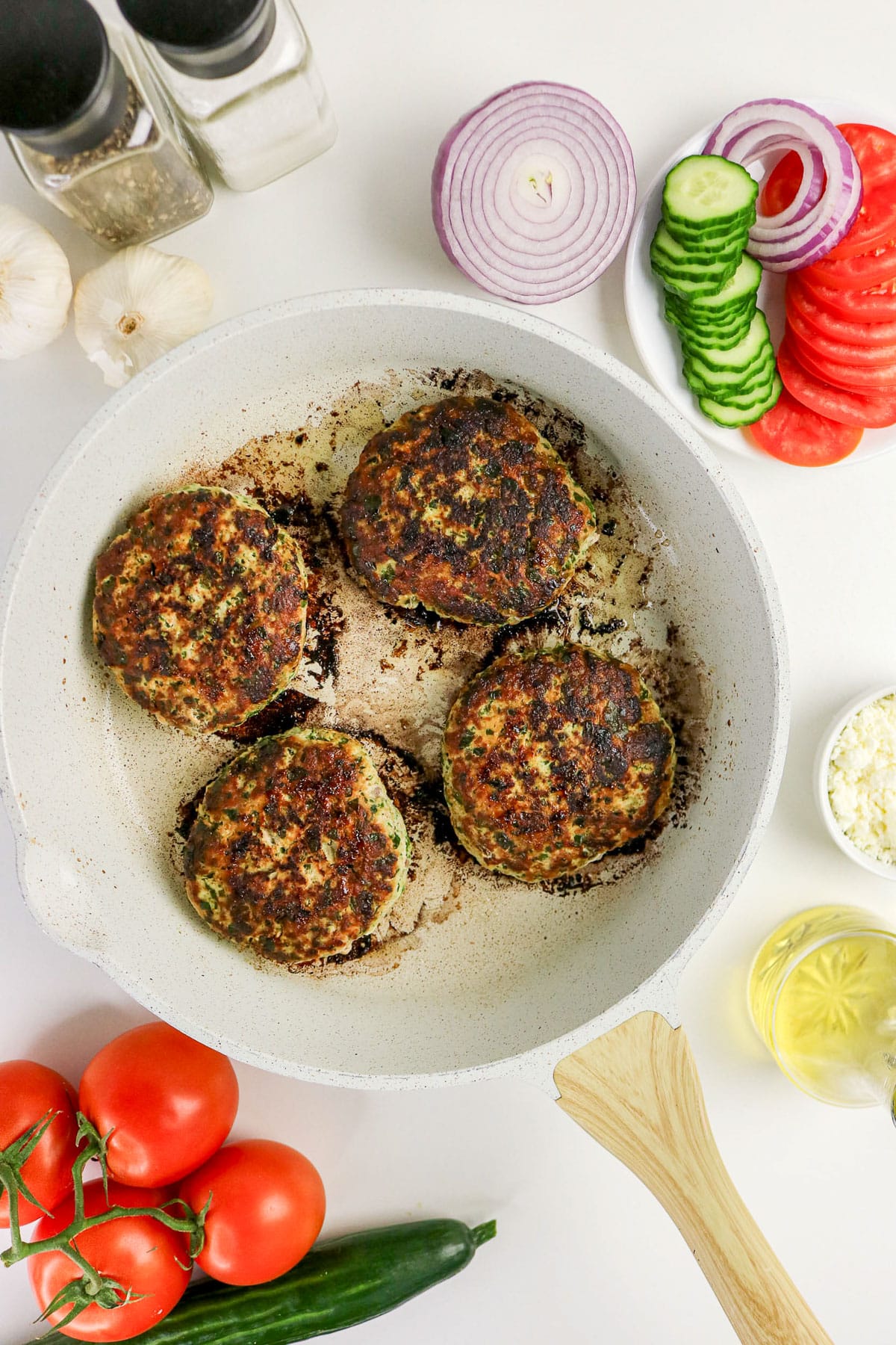 Four browned patties cooking in a white pan, surrounded by fresh veggies and seasonings on a white surface.