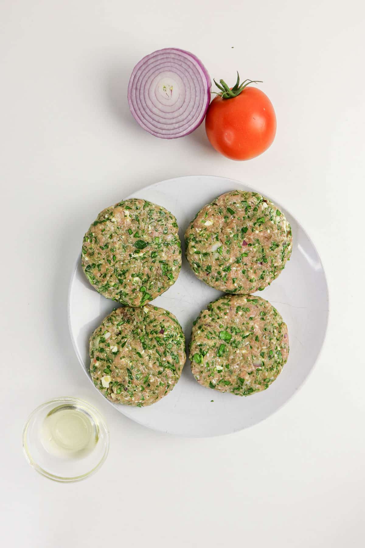Four uncooked patties on a plate, with a red onion half, a tomato, and a small bowl of oil nearby.