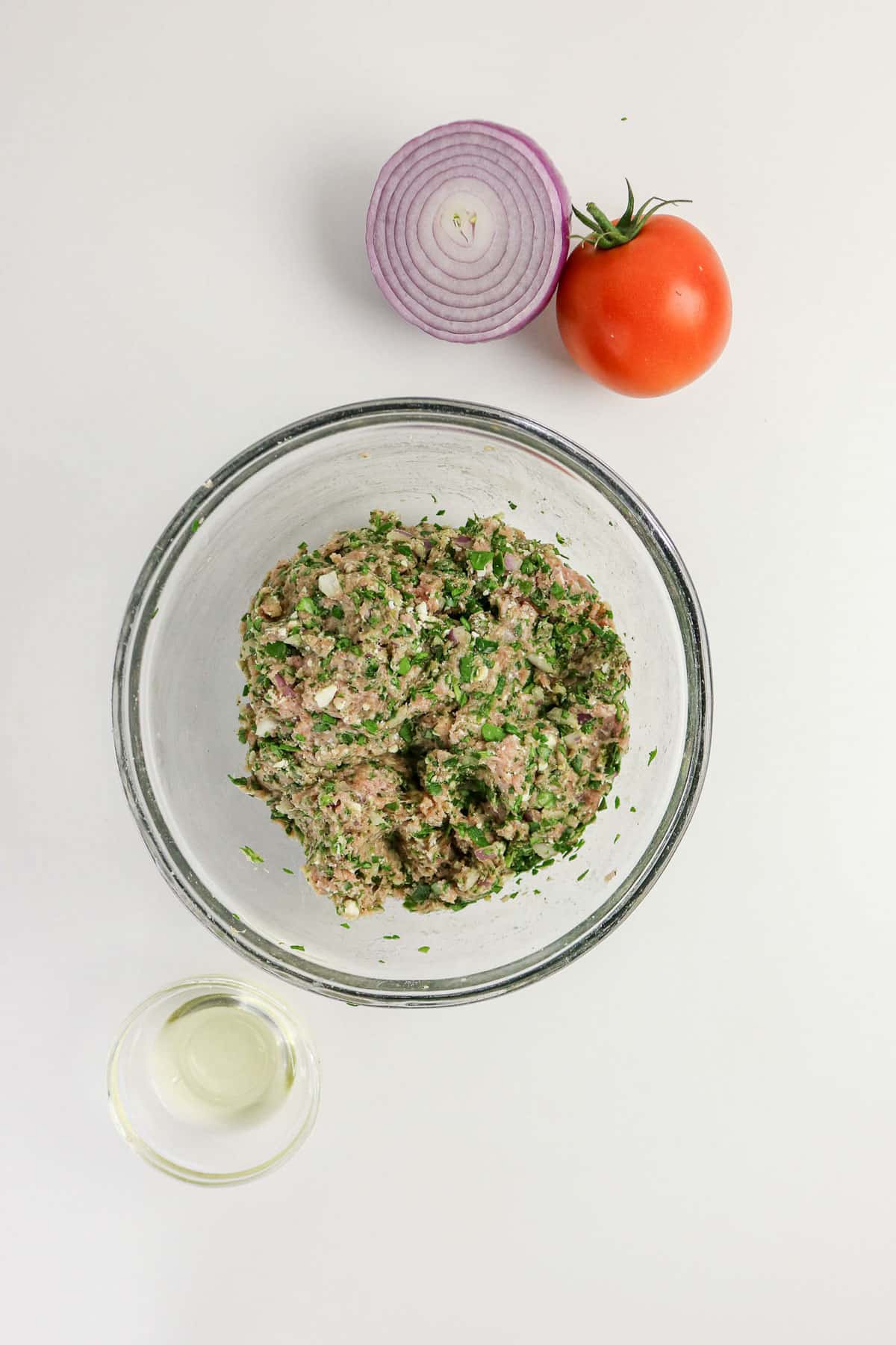 A bowl of seasoned ground meat with parsley, next to half a red onion, a tomato, and a small cup of liquid.