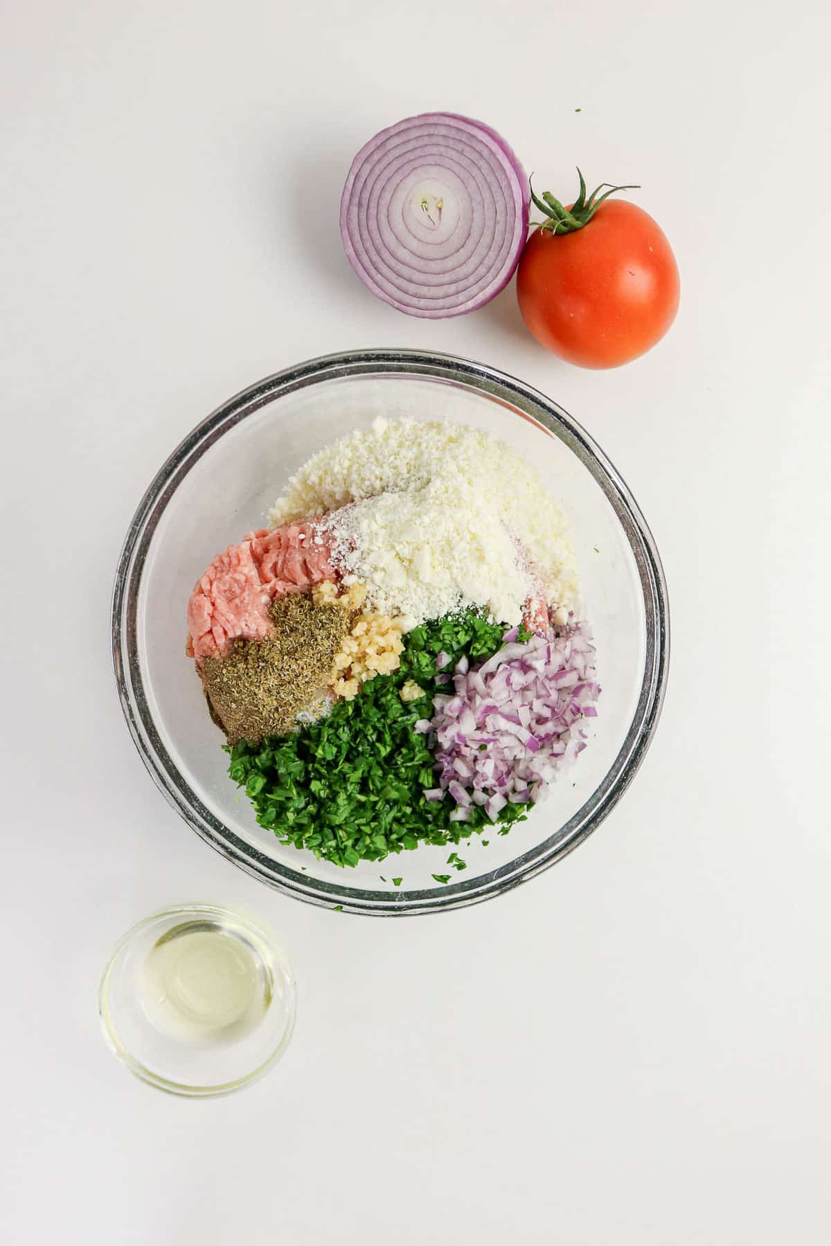 A bowl with ground turkey, chopped onion, herbs, spices, and cheese, with a tomato and half red onion nearby.