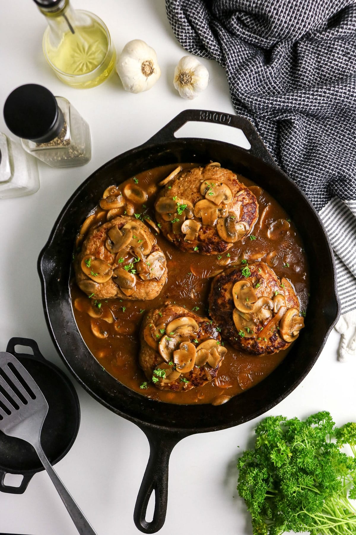 Three Salisbury steaks with mushroom gravy in a cast iron skillet, surrounded by seasonings and fresh herbs.