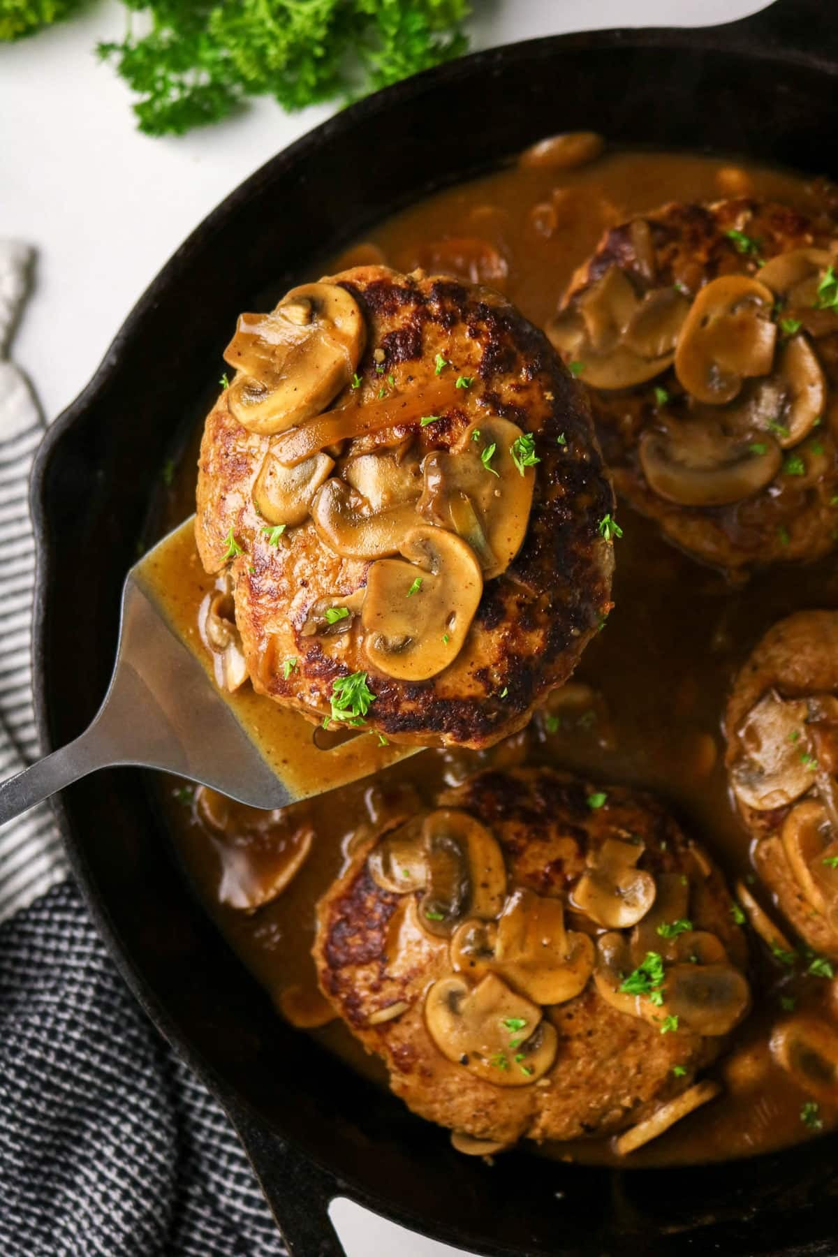 A spatula lifts a Salisbury steak topped with mushrooms and gravy from a skillet with more steaks inside.