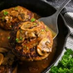 A Salisbury steak topped with mushrooms and gravy in a skillet, being lifted with a spatula.