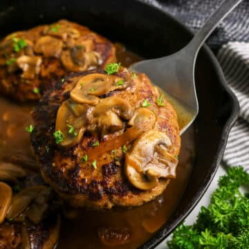 A Salisbury steak topped with mushrooms and gravy in a skillet, being lifted with a spatula.