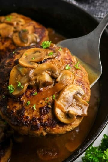 A Salisbury steak topped with mushrooms and gravy in a skillet, being lifted with a spatula.