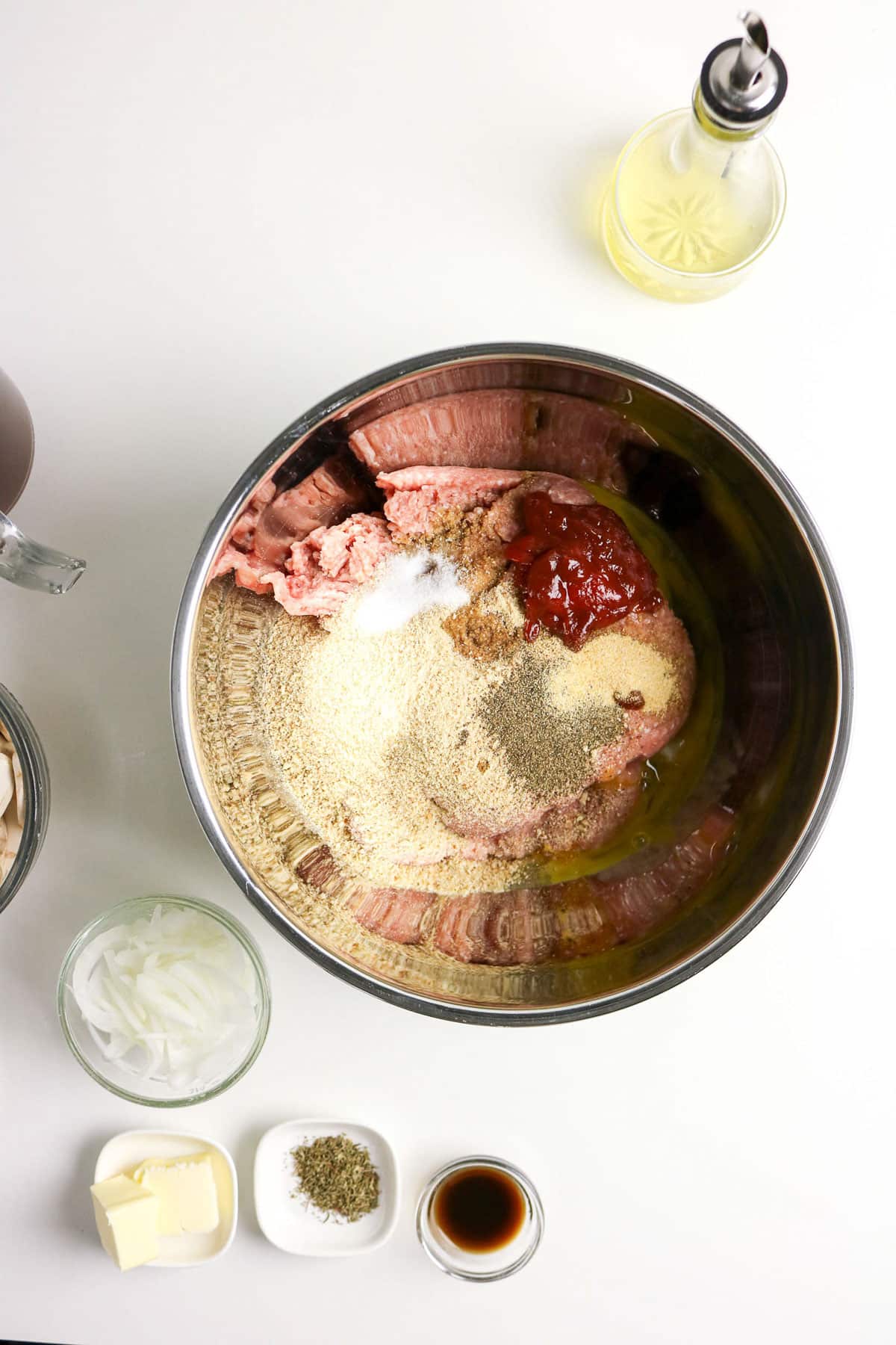 Large bowl with ground meat, ketchup, breadcrumbs, egg, and seasonings; surrounding bowls with onions, butter, and more.