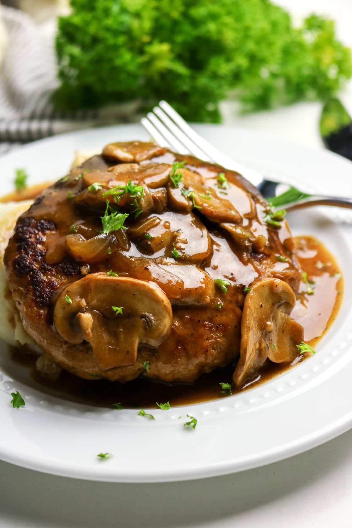 A Salisbury steak topped with mushroom gravy, garnished with parsley, on a white plate with a fork.