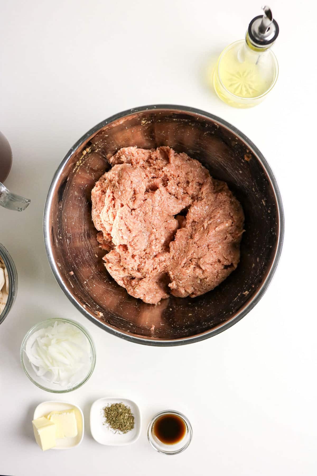 Large metal bowl of ground meat, surrounded by oil, onions, butter, seasonings, and sauce on a white surface.
