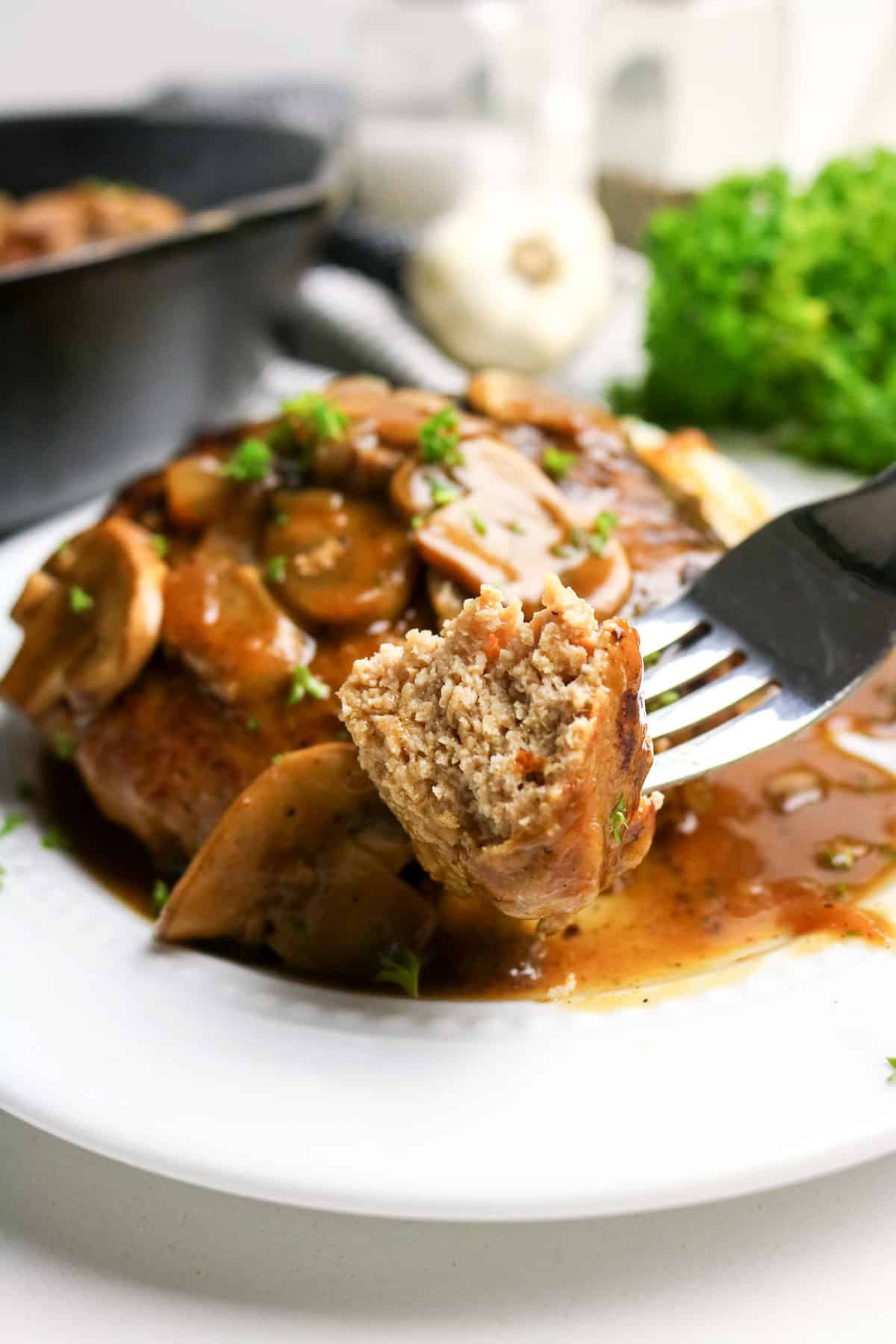 A fork holds a bite of Salisbury steak with mushrooms and gravy, served on a white plate.