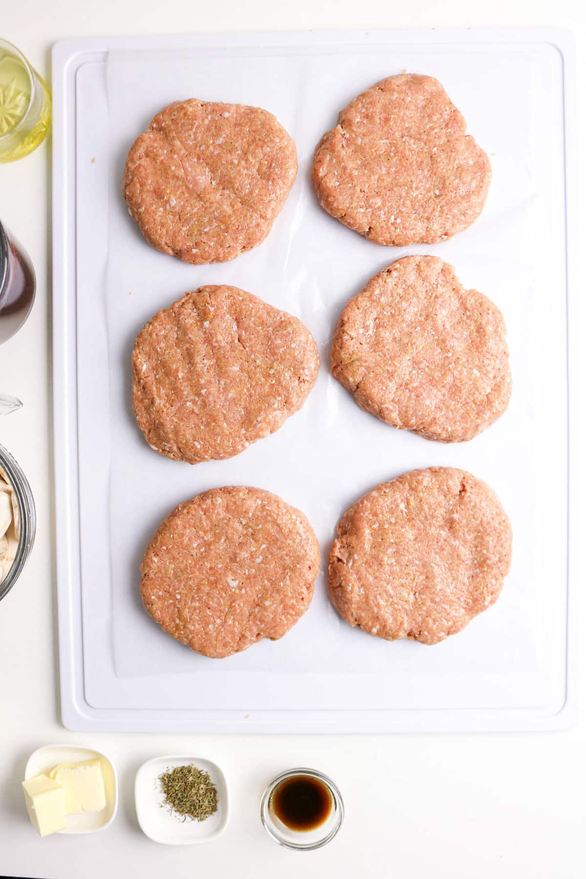 Six raw turkey burger patties on a white tray with seasonings, butter, and sauce in small dishes beside them.
