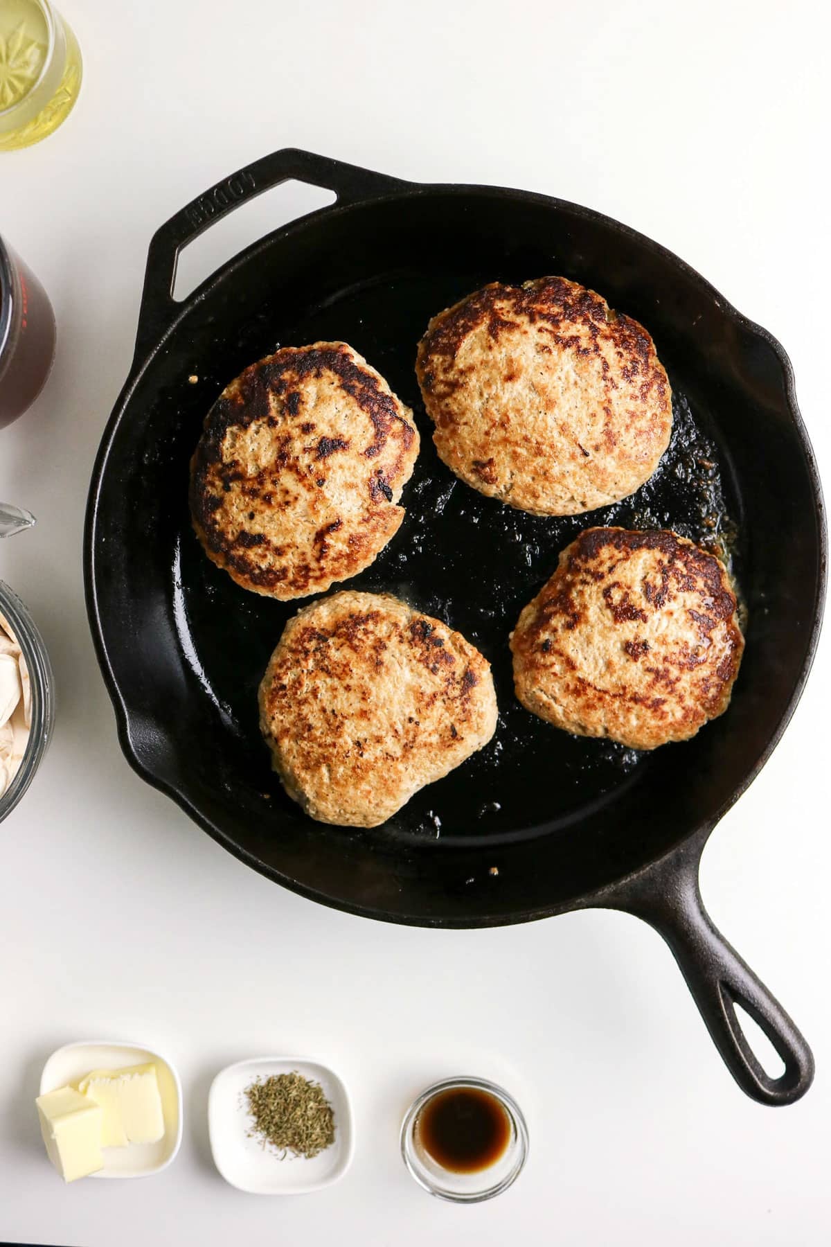 Four browned patties are cooking in a black cast iron skillet, surrounded by small bowls of ingredients.