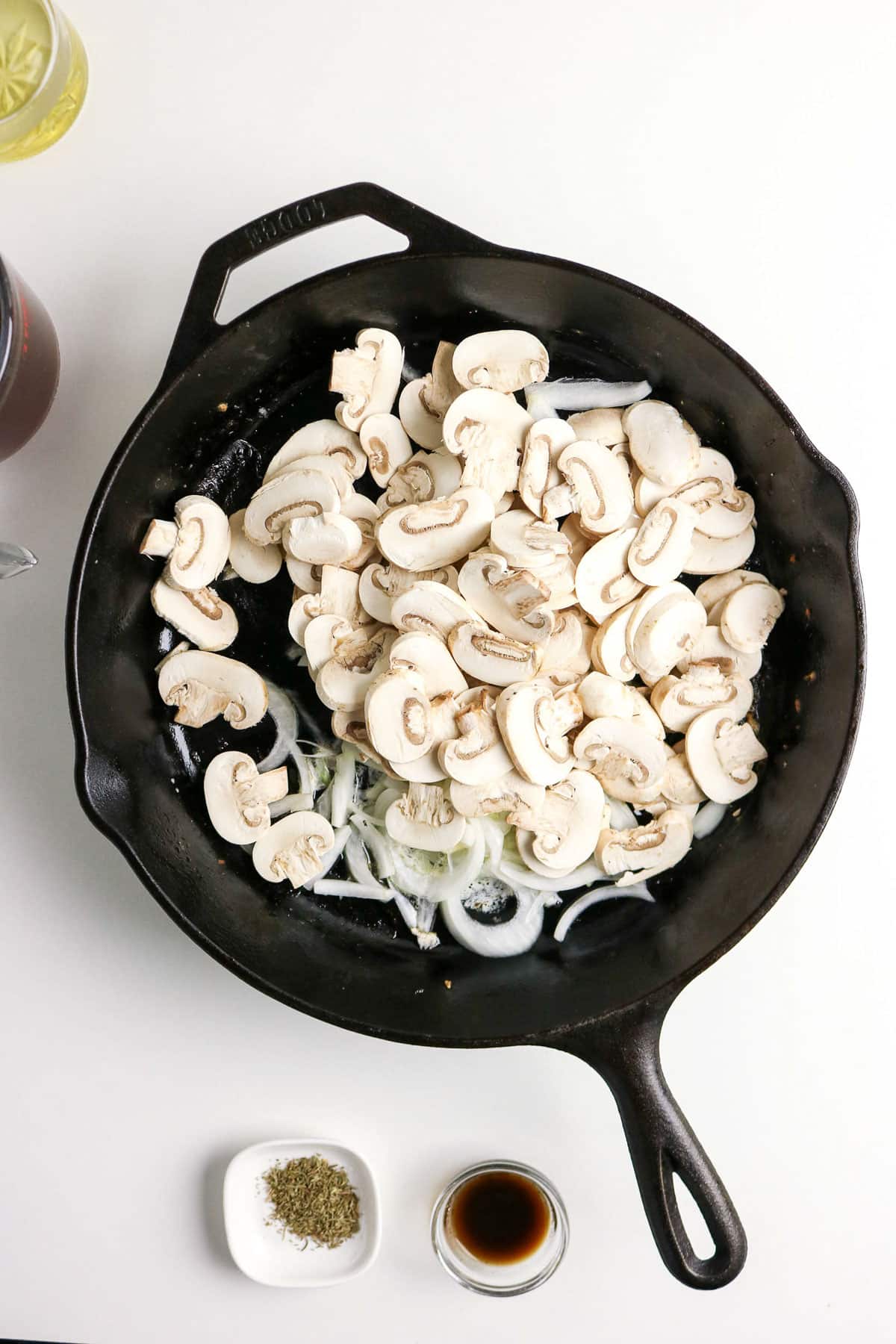 Sliced mushrooms and onions in a black skillet, with herbs and sauce in small bowls nearby on a white surface.