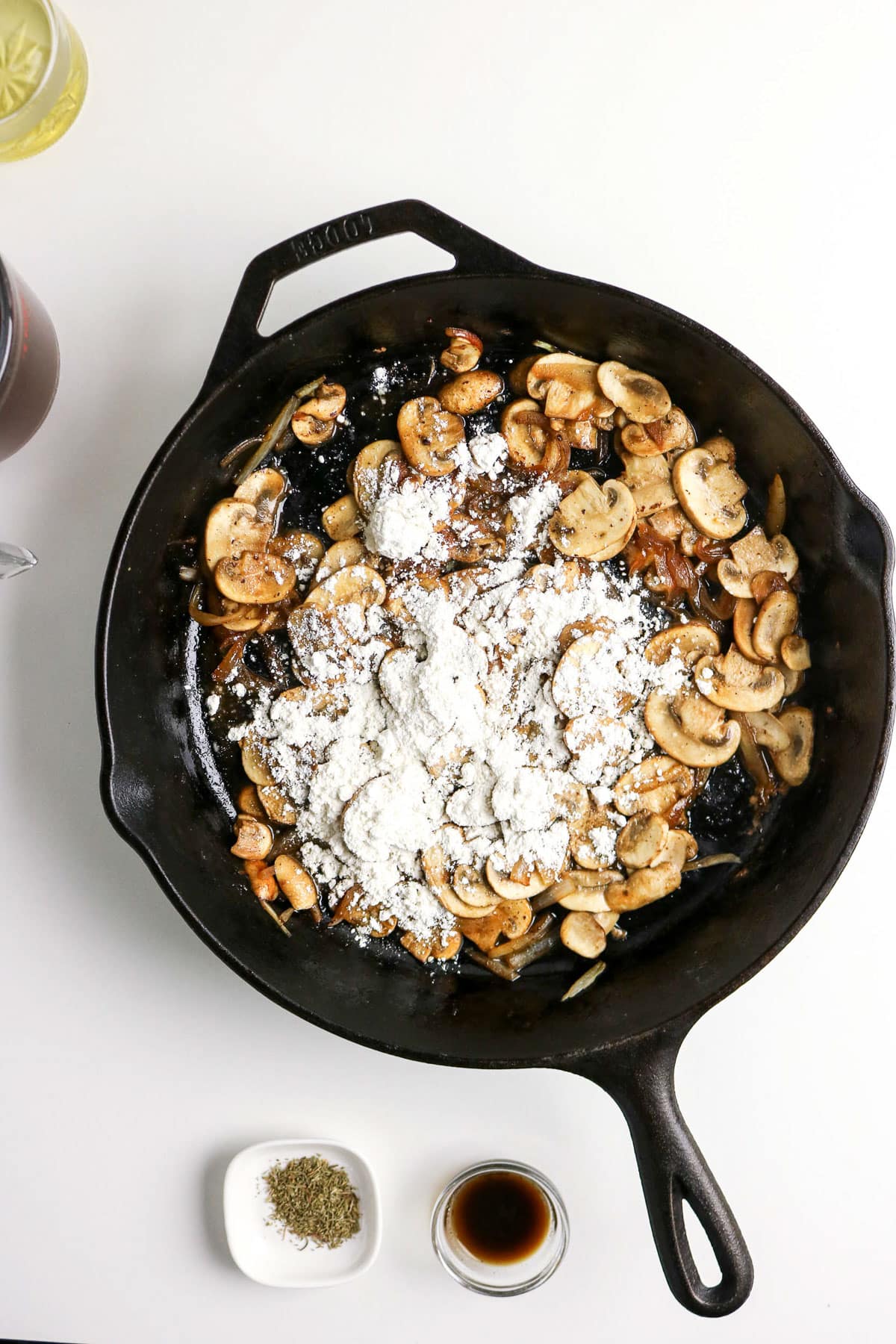 Sliced mushrooms and flour in a cast iron skillet, with seasonings and sauce in small bowls nearby on a white surface.