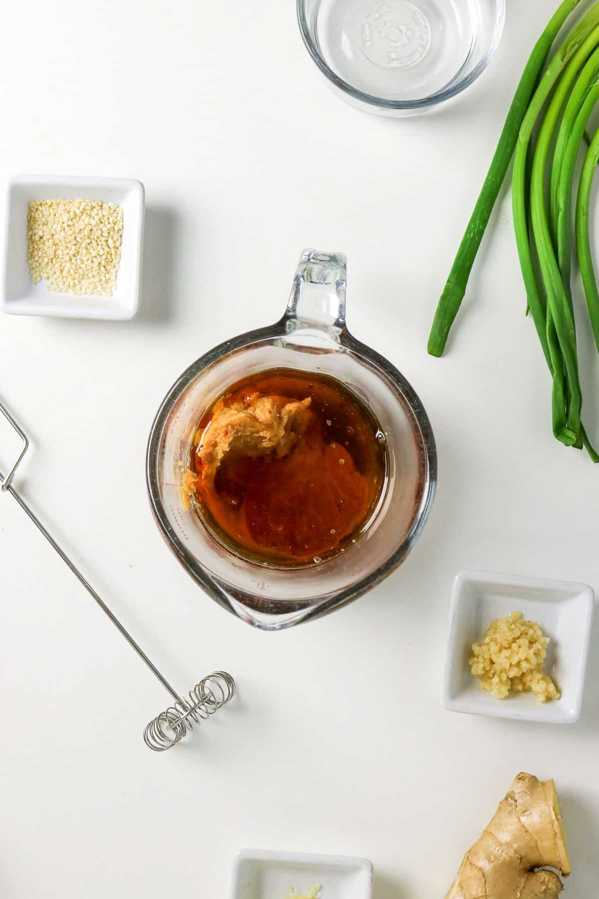 Glass measuring cup with sauce ingredients, surrounded by sesame seeds, green onions, ginger, garlic, and water.