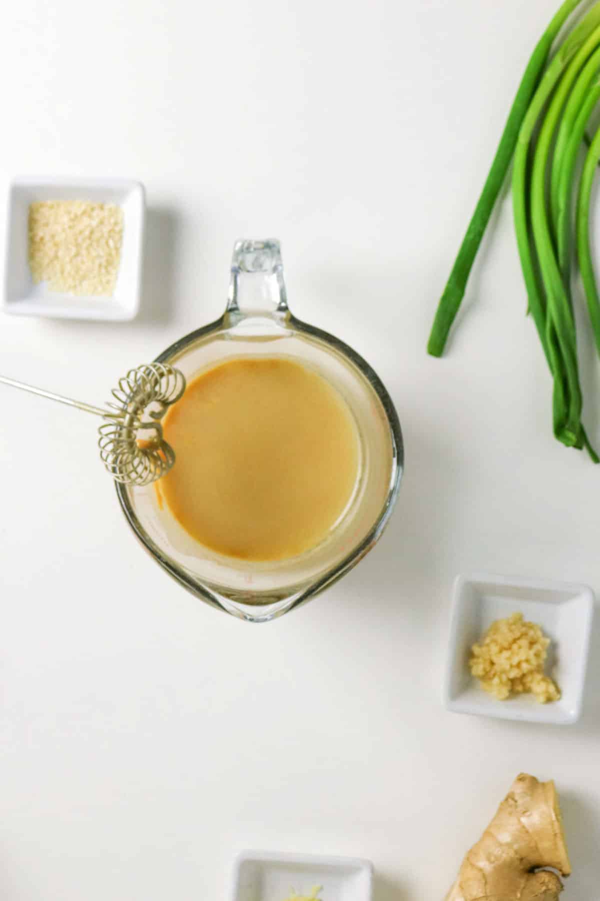 Glass measuring cup of creamy dressing with a whisk, surrounded by green onions, and small bowls of ingredients.