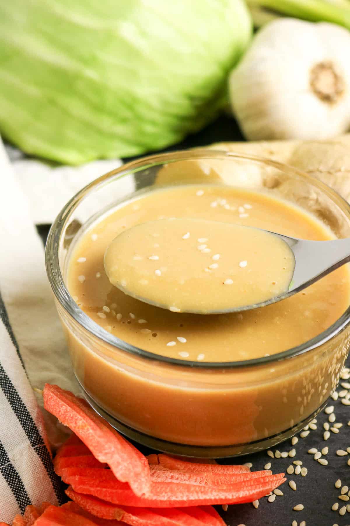 A bowl of creamy sesame dressing with a spoon, surrounded by cabbage, garlic, ginger, and carrot slices.