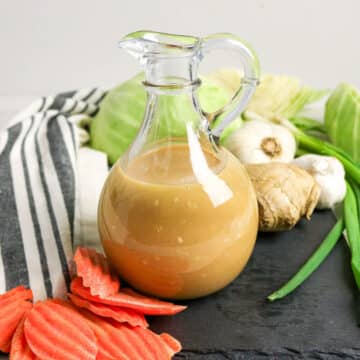 A glass jug of brown dressing with ginger, garlic, cabbage, green onion, and sliced carrots around it.