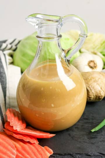 A glass jug of brown dressing with ginger, garlic, cabbage, green onion, and sliced carrots around it.