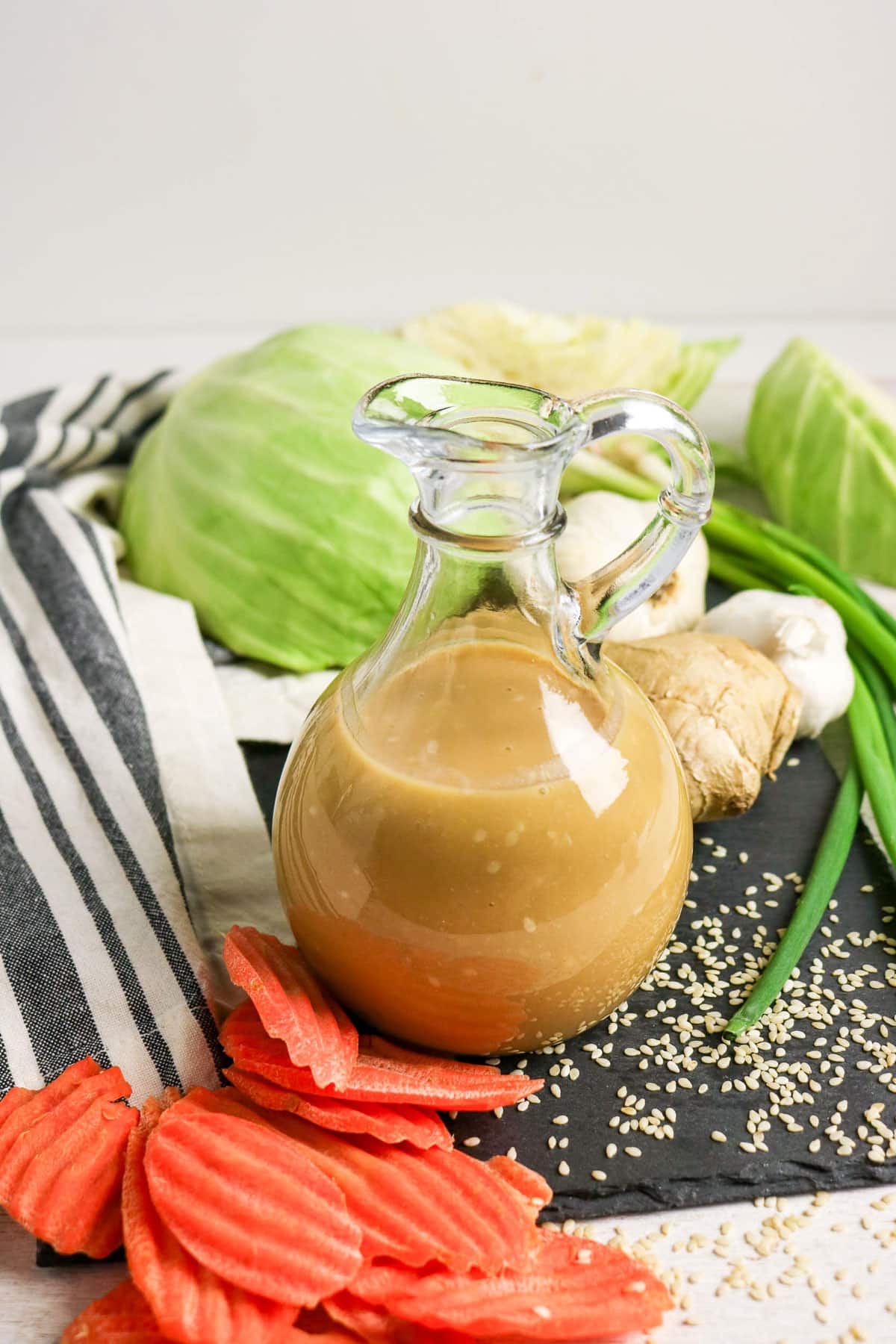 A glass pitcher of dressing with cabbage, carrots, ginger, green onions, garlic, and sesame seeds nearby.