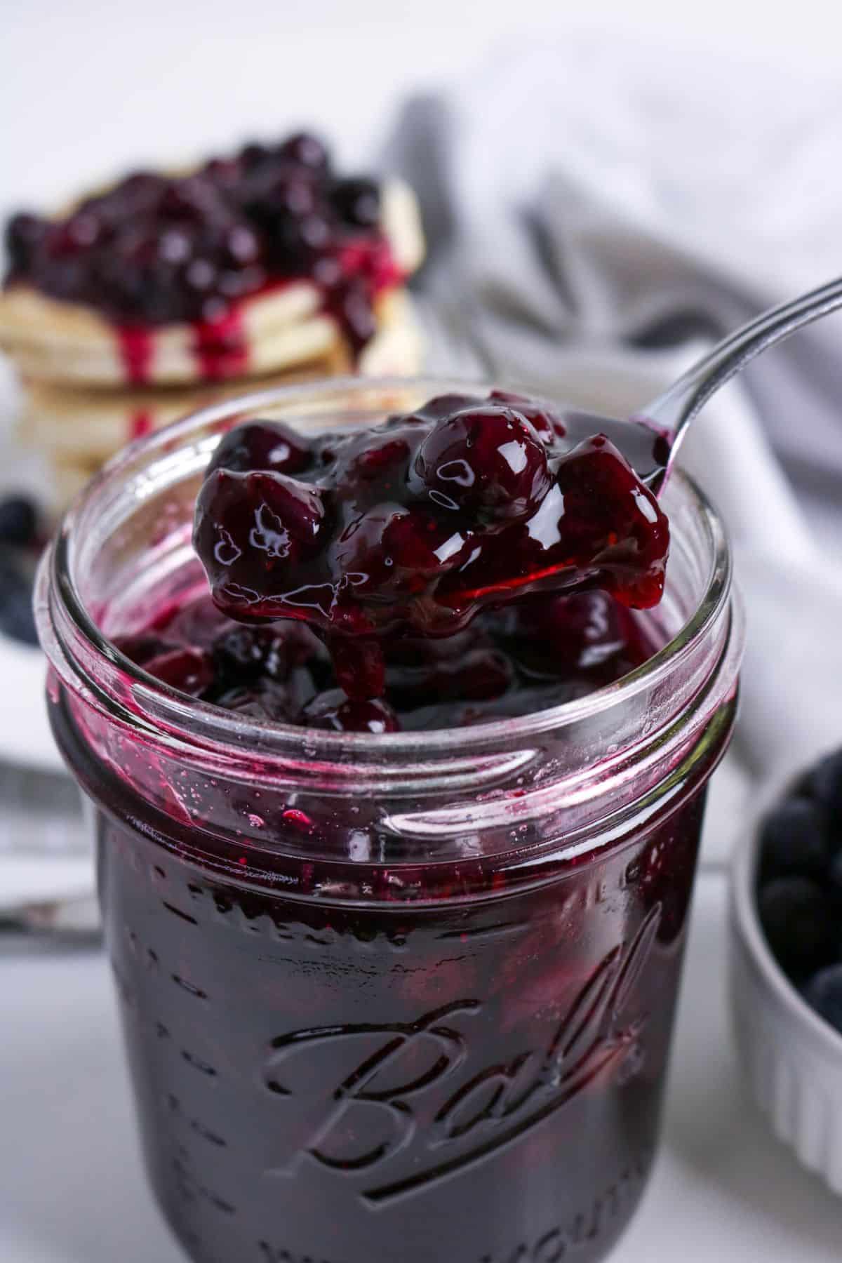 A spoonful of blueberry jam being lifted from a glass jar, with pancakes and blueberries in the background.