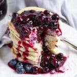 A stack of pancakes topped with blueberry sauce and fresh blueberries on a white plate with a fork.