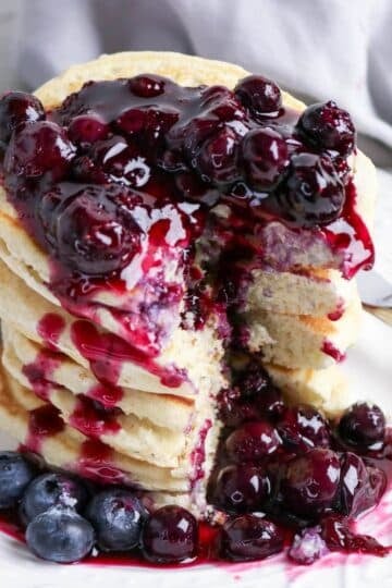 A stack of pancakes topped with blueberry sauce and fresh blueberries on a white plate with a fork.