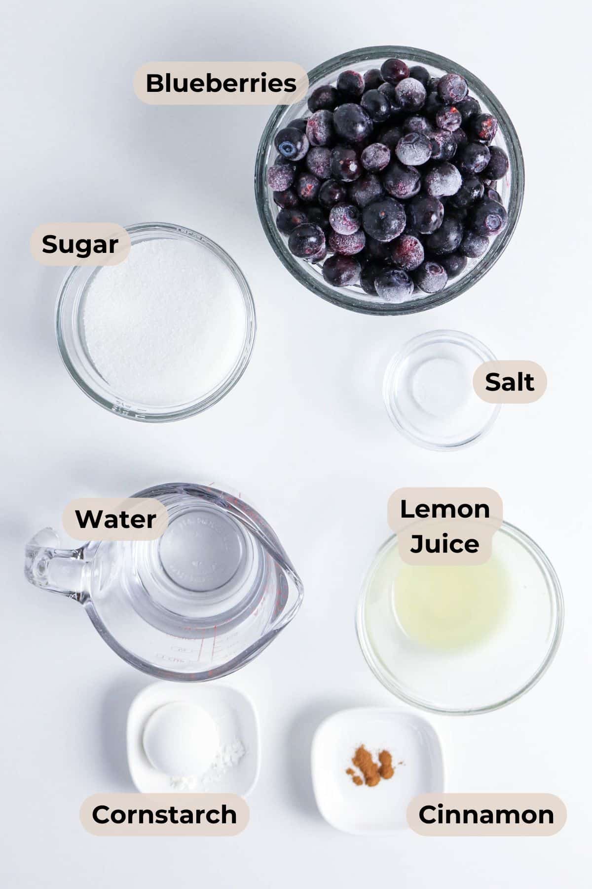 Bowls of blueberries, sugar, salt, lemon juice, cornstarch, cinnamon, and a measuring cup of water on a white surface.