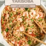A skillet of shrimp yakisoba with ramen noodles, vegetables, and green onions, with wooden tongs.