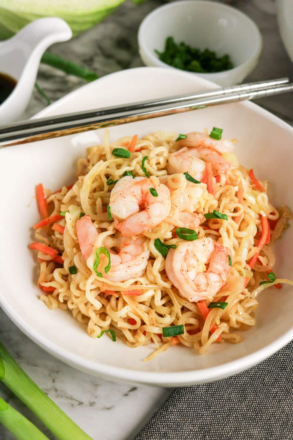 A bowl of shrimp stir-fry noodles with carrots, green onions, and chopsticks on the edge of the bowl.