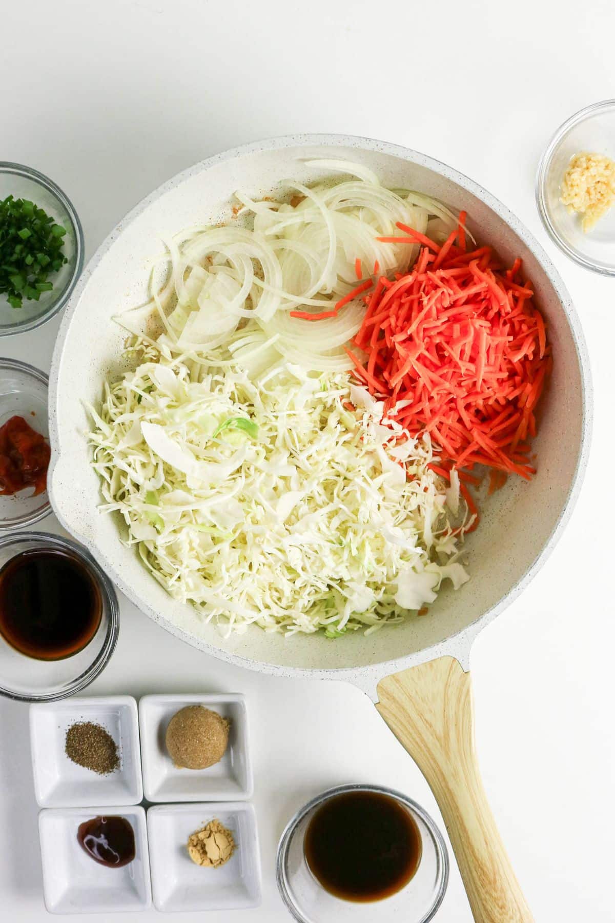 A skillet with sliced onions, shredded carrots, and cabbage, surrounded by bowls of sauces and seasonings.