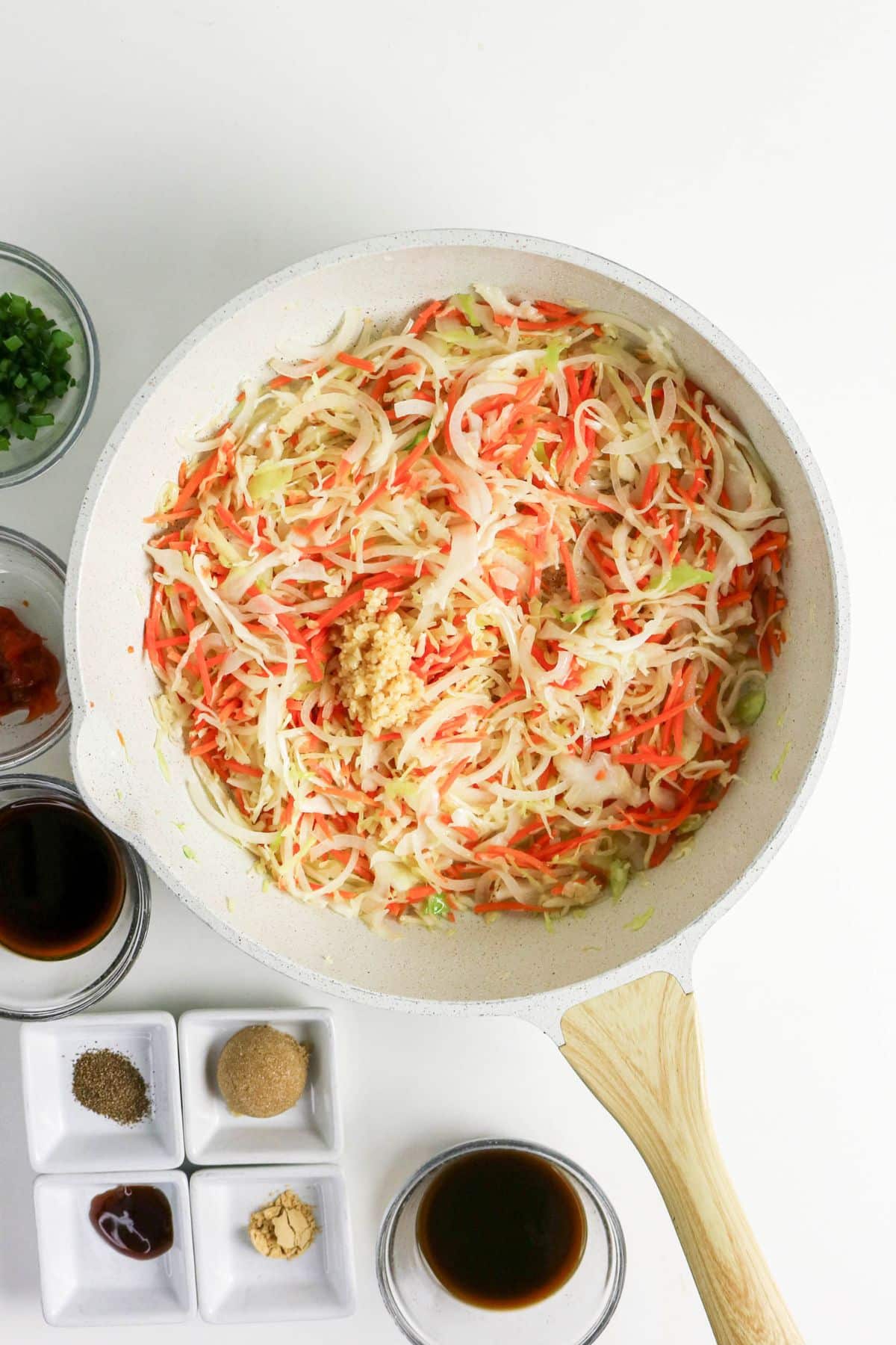 Top-down view of shredded cabbage and carrots in a white pan, surrounded by small bowls of sauces and spices.